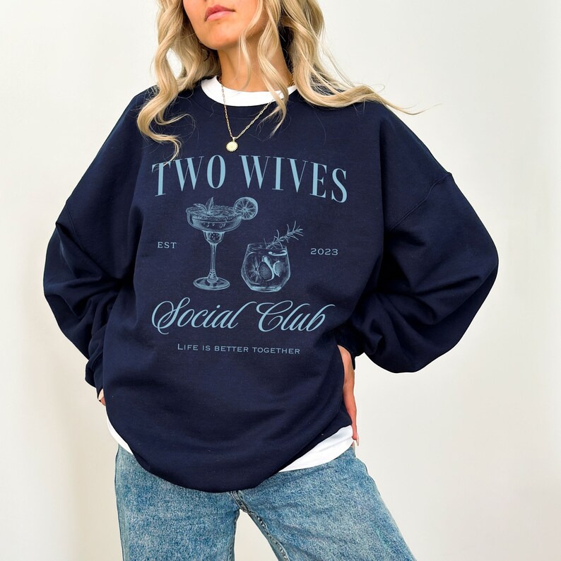 Puede incluir: Sudadera azul marino con el texto "TWO WIVES SOCIAL CLUB" en azul claro. El dise&ntilde;o incluye ilustraciones de dos copas de c&oacute;ctel y la frase "LIFE IS BETTER TOGETHER". La sudadera tiene cuello redondo blanco.