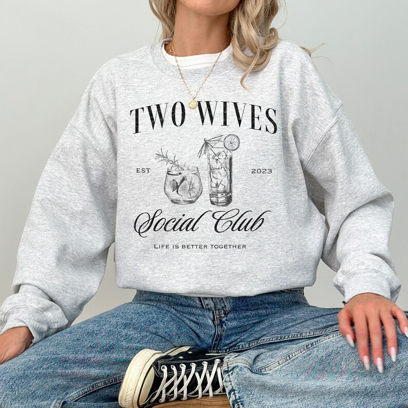 Puede incluir: Una sudadera gris claro con el texto "TWO WIVES Social Club" en escritura negra. El dise&ntilde;o incluye ilustraciones de dos c&oacute;cteles y la frase "LIFE IS BETTER TOGETHER". La sudadera se lleva con vaqueros azules y zapatillas negras.
