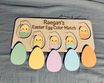 May include: Wooden Easter egg color matching game with the name "Raegan's Easter Egg Color Match" engraved on the top. The game features four chicks in eggs with different colored shells and four blank egg shapes to match.