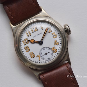 Op de afbeelding: Een zilveren polshorloge met een witte wijzerplaat en oranje cijfers. Het horloge heeft een bruin leren bandje en staat "Waltham" op de wijzerplaat.