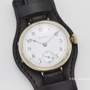 以下が含まれることがあります： 白い文字盤、黒い数字、サブダイヤルを備えたヴィンテージのハミルトン腕時計。 金色のベゼルと黒い革ストラップが付いています。 時計は黒い革のカフにセットされています。