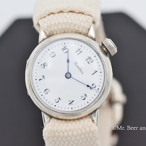 May include: A vintage Hamilton wristwatch with a white face, black numerals, and blue hands. The watch has a silver-colored case and a cream-colored canvas band. The watch is set against a dark background.