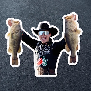 May include: A sticker featuring a person wearing a black cowboy hat and sunglasses, holding up two large fish. The person is smiling. The sticker has a white border and is set against a dark gray background.