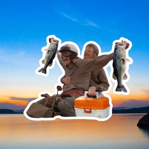 May include: A person wearing a helmet and holding two fish, sitting on a small boat with a tackle box. The scene is set against a sunset over a lake.