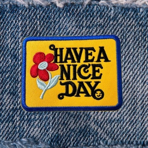 May include: Yellow embroidered patch with a blue border. The patch features a red flower and the text "HAVE A NICE DAY" in black.