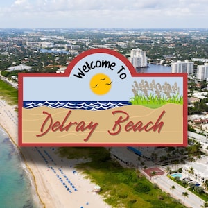 May include: An aerial view of Delray Beach, Florida, with a welcome sign that reads "Welcome to Delray Beach". The sign is red, white, and blue with a beach scene and a yellow sun.
