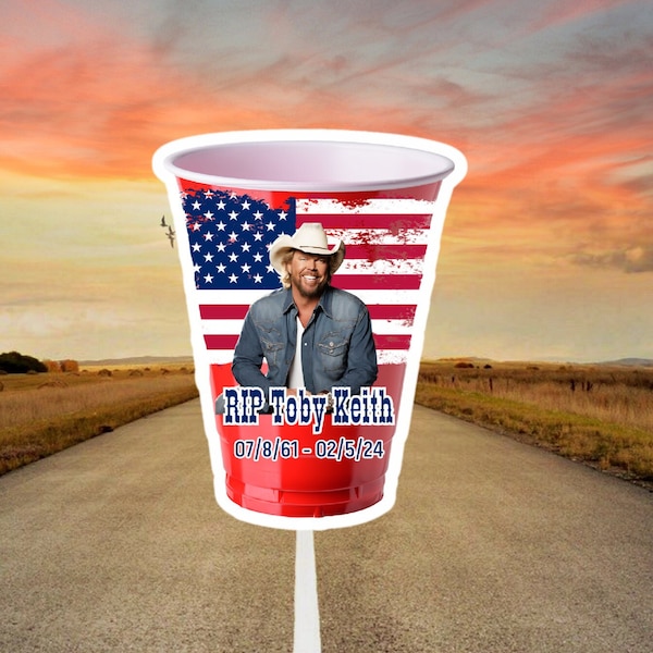May include: Red plastic cup featuring an American flag design and a picture of Toby Keith. The text "RIP Toby Keith 07/8/61 - 02/5/24" is printed on the cup.