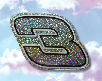 May include: A silver glitter sticker with a black outline of the letter "B" against a background of pink and blue clouds.