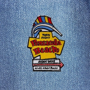 May include: A yellow and red embroidered patch with the text "Pensacola Beach" and "Turn Right" on a blue denim background. The patch also includes the text "Scenic Drive" and "World's Whitest Beaches".