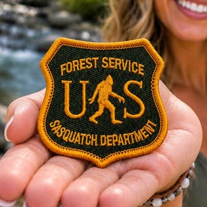 May include: A shield-shaped embroidered patch with a dark green background and gold border. The text reads FOREST SERVICE and SASQUATCH DEPARTMENT, with the letters U and S flanking a gold silhouette of a Sasquatch in the center of the patch.