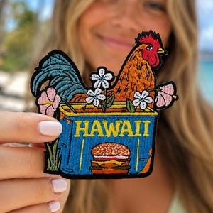 May include: An embroidered patch featuring a rooster with orange feathers and a blue tail, sitting in a blue container labeled HAWAII. The design includes white and pink hibiscus flowers and a cheeseburger graphic on the front of the container.