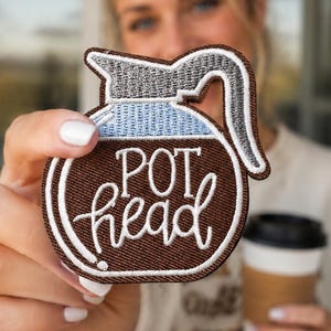 May include: A coffee pot shaped embroidered patch with a brown base, light blue middle section, and grey handle. The text POT head is stitched in white thread across the center of the patch, which features a dark brown border outline.