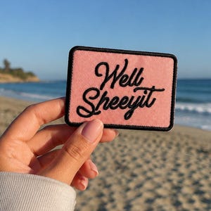 May include: A rectangular patch with a pink background and black border. The patch features the phrase "Well Sheeyit" in a cursive font. The patch is held up against a beach backdrop with the ocean and sky in the background.