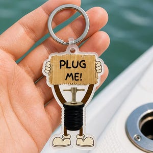 May include: A novelty keychain featuring a cartoon-style illustration of a black and brown plug holding a sign that reads "PLUG ME!". The keychain has a silver metal ring and is held against a blurred background.