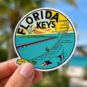 May include: A circular sticker featuring a map of the Florida Keys. The sticker has a yellow border with the words "Florida Keys" in black. The map shows the islands, with illustrations of a bird, turtle, and marine life.