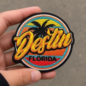 May include: Embroidered patch featuring the word "Destin" in a yellow script font, set against a sunset design with a palm tree silhouette. The word "FLORIDA" is at the bottom. The patch has a black border.