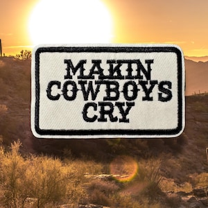 May include: Embroidered patch with the text "MAKIN COWBOYS CRY" in black lettering on a white background, framed by a black border. The patch is set against a desert sunset backdrop.