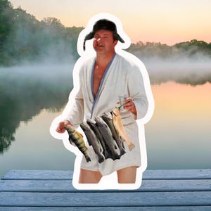 May include: A man wearing a white bathrobe and a black fur hat holds a string of fish in his hands. The man is standing on a wooden dock with a lake in the background.