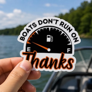 May include: A white sticker with the text "BOATS DON'T RUN ON Thanks" in a black and orange design. The sticker features a fuel gauge graphic. The sticker is held by a hand against a blurred background of water and trees.