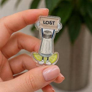 May include: A clear acrylic pin featuring a cartoon salt shaker character holding a sign that reads "LOST." The salt shaker is standing on a lime slice and has small white shoes. The pin is held by a person.