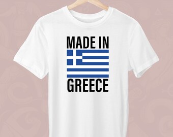 Peut inclure: T-shirt blanc avec l'inscription "MADE IN GREECE" en noir. Le drapeau grec, avec ses rayures bleues et blanches et son carré bleu, est imprimé sur le devant. Le t-shirt est suspendu à un cintre en bois.