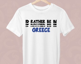Peut inclure: T-shirt blanc avec l'inscription "I'D RATHER BE IN GREECE" en noir et bleu. Le t-shirt est suspendu à un cintre en bois. Le texte est centré sur le devant du t-shirt.