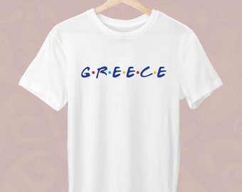 Peut inclure: T-shirt blanc avec le mot "GREECE" en lettres bleues, jaunes, rouges et bleues. Le design rappelle la série télévisée "Friends". Le t-shirt est suspendu à un cintre en bois.