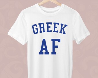 Peut inclure: T-shirt blanc avec l'inscription "GREEK AF" en bleu, présenté sur un cintre en bois. Le texte est en gras, dans une police de style universitaire. Le t-shirt est fait d'une matière douce et légère.