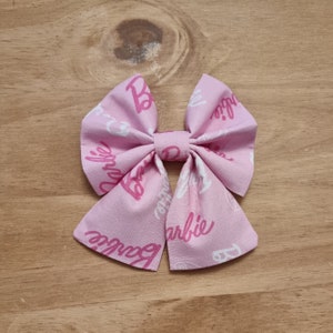 Peut inclure: Noeud en tissu rose avec un imprimé logo Barbie blanc et rose.
