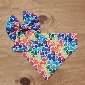 May include: A colorful pet accessory set featuring a rainbow heart pattern. The set includes a bow tie and a triangular bandana. The hearts are in shades of red, orange, yellow, green, blue, and purple, set against a white background.