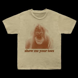 May include: Beige t-shirt featuring a brown orangutan graphic. The text "show me your toes" is printed in brown below the image. The shirt has a crew neck and short sleeves.