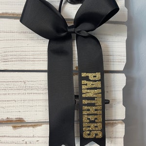 May include: Black hair bow with a gold glitter "Cheerleaders" text.