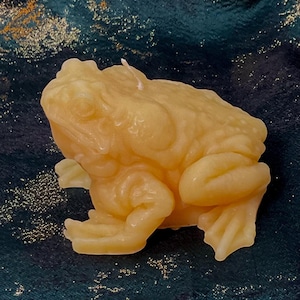 May include: A light yellow frog-shaped candle. The candle has a detailed texture, with visible bumps and folds, and is positioned on a dark teal fabric with gold accents. The candle is a decorative item.