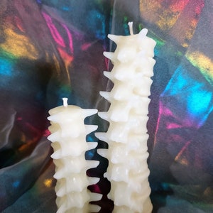 May include: Two white, spine-shaped candles, one taller than the other, on a colorful iridescent background.