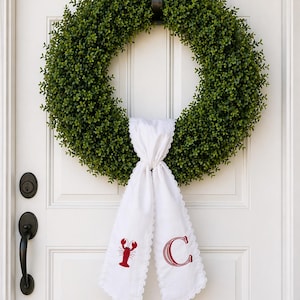 May include: A circular green wreath made of foliage hangs on a white door. A white, scalloped-edged fabric is tied to the wreath, with a red embroidered lobster and the letter "C". The door has black hardware.