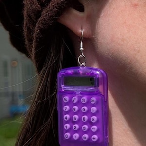 May include: Purple calculator earrings. The earrings are shaped like a small calculator with a clear plastic case and silver metal hooks.