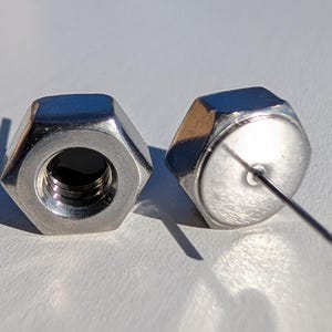 May include: Two shiny, hexagonal metal objects. One has a threaded hole, the other has a thin, straight metal rod extending from its center. The objects are on a light-colored surface, casting shadows.