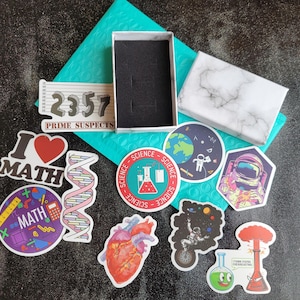 May include: A collection of colorful stickers featuring science and math themes. The stickers include a beaker, a heart, a DNA strand, a rocket ship, a planet, and a math equation. The stickers are on a blue background with a white marble box and a white box with the text "2357 Prime Suspects".