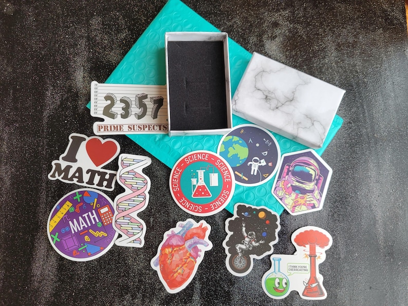 May include: A collection of colorful stickers featuring science and math themes. The stickers include a beaker, a heart, a DNA strand, a rocket ship, a planet, and a math equation. The stickers are on a blue background with a white marble box and a white box with the text "2357 Prime Suspects".