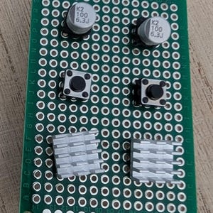 May include: A rectangular green printed circuit board featuring a grid of through-holes, two cylindrical silver capacitors labeled K2 100 6.3J, two black square tactile push-button switches, and two silver-colored square aluminum heat sinks.