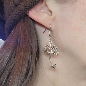 Op de afbeelding: Zilveren oorbellen in de vorm van een neuron, een type zenuwcel. De oorbellen hangen aan het oor van een persoon.