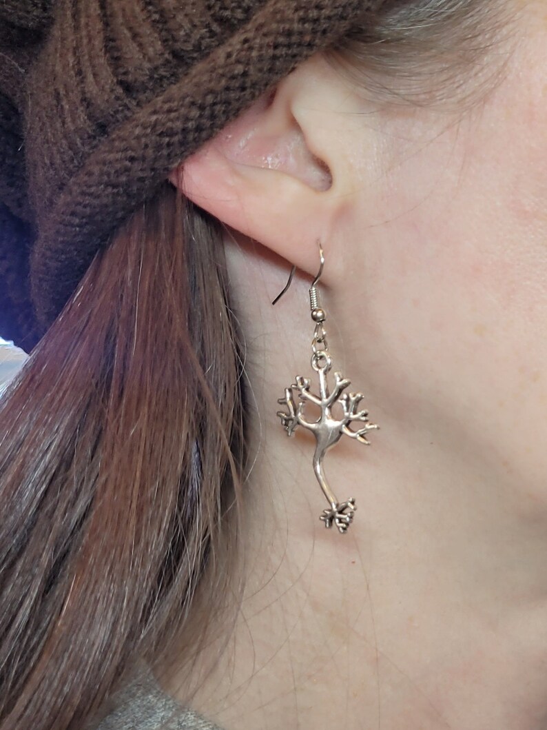 Op de afbeelding: Zilveren oorbellen in de vorm van een neuron, een type zenuwcel. De oorbellen hangen aan het oor van een persoon.