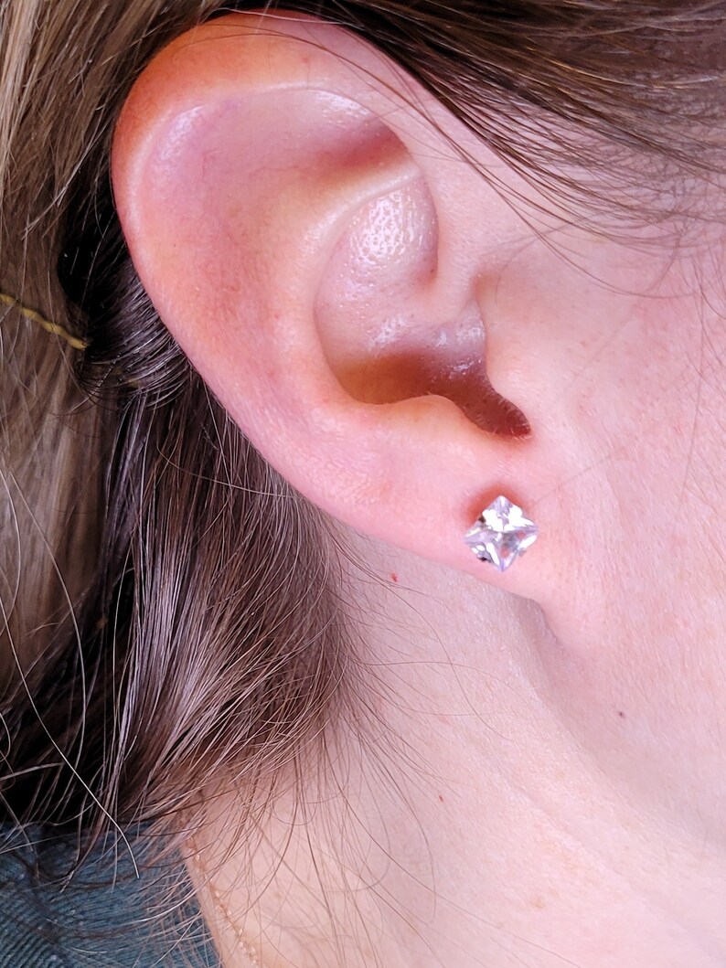 Op de afbeelding: Een close-up van iemands oor met een kleine, vierkante, doorzichtige oorbel.