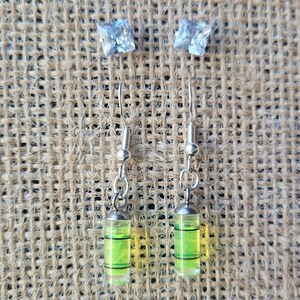 May include: A pair of silver earrings with a green and yellow level bubble charm. The earrings have a clear crystal at the top.