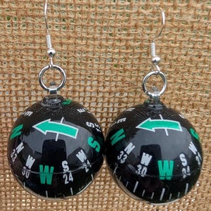 May include: A pair of novelty earrings featuring black and green compass designs. Each earring has a spherical shape with directional markings and a green arrow. The earrings hang from silver-colored hooks.