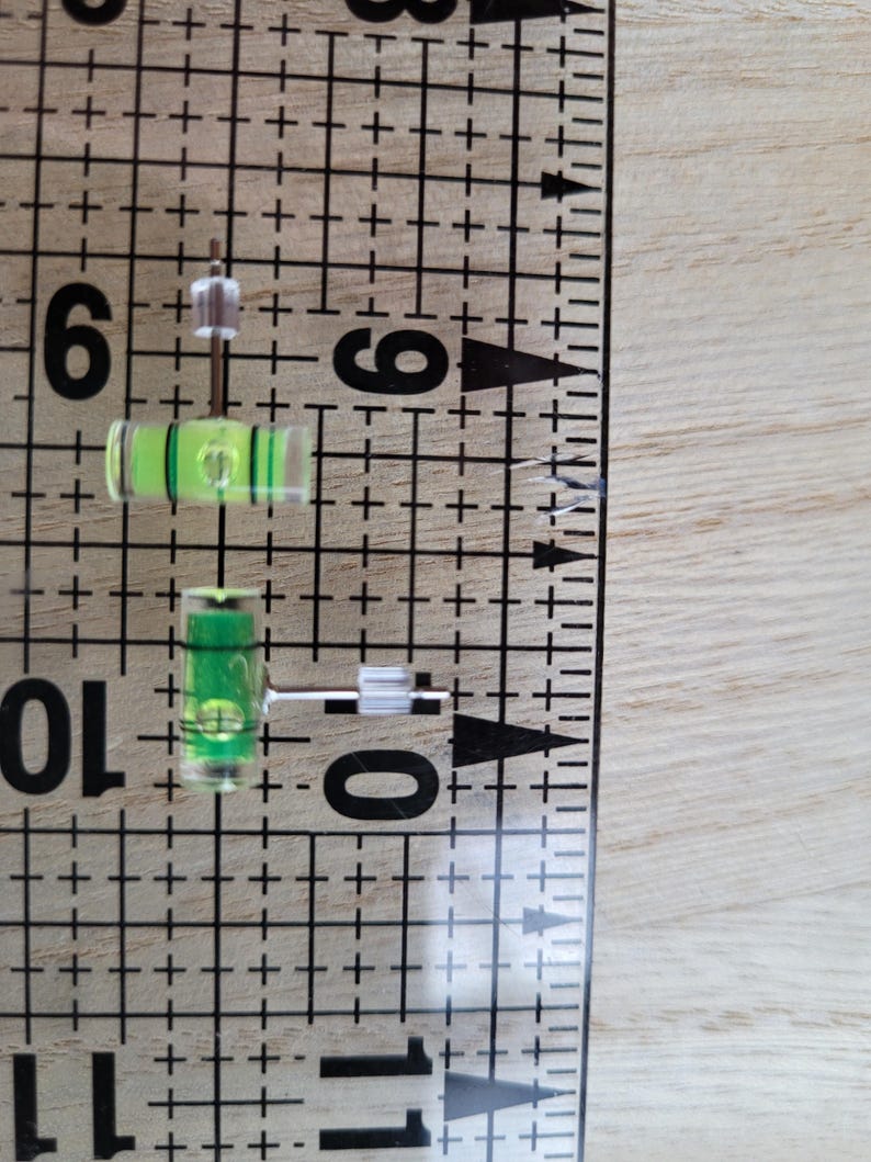 May include: Two small green and clear levelers with a white plastic base. The levelers are about 1 inch long and are sitting on a white grid.