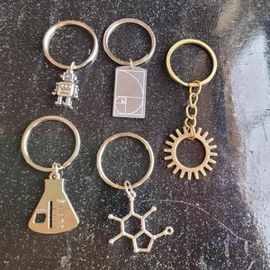 May include: Five silver keychains with different charms. The charms include a robot, a beaker, a molecule, a sun, and a square with a spiral pattern. The keychains are on a black background.