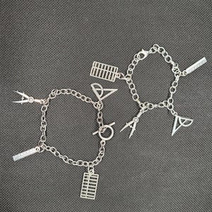 May include: Two silver-toned charm bracelets with various school-themed charms, including an abacus, protractor, ruler, and compass. The bracelets are displayed on a dark gray surface. The charms are detailed and add a unique touch.