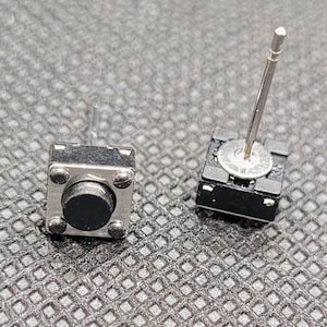 May include: Two small black push button switches with silver metal contacts and a silver metal pin on top. The switches are on a gray textured surface.