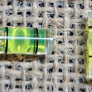 May include: Two clear plastic spirit levels with green liquid and black lines. The levels are on a brown burlap background.